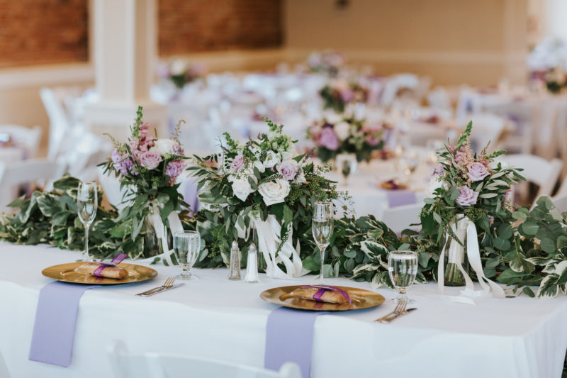 Bride & Groom's sweetheart table with gold chargers, lavender napkins and lush greenery garlands