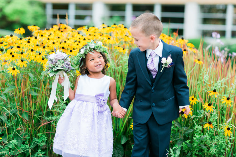Ring Bearer and Flower Girl holding hands in front of a patch of yellow flowers wearing flower crown and purple striped tie